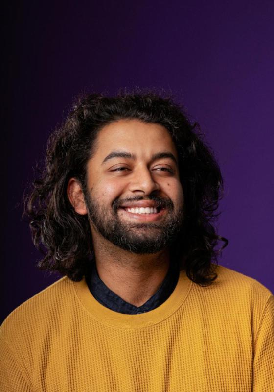 Portrait of a smiling man with curly hair wearing a yellow sweater, against a purple background.