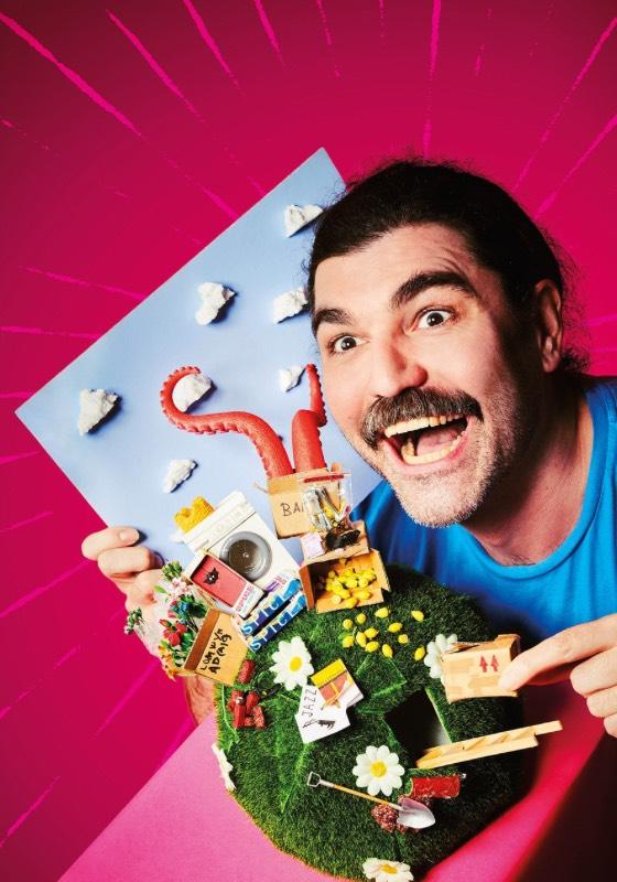 John Luke Roberts holds up a grass covered globe and points to miniatures of his show.