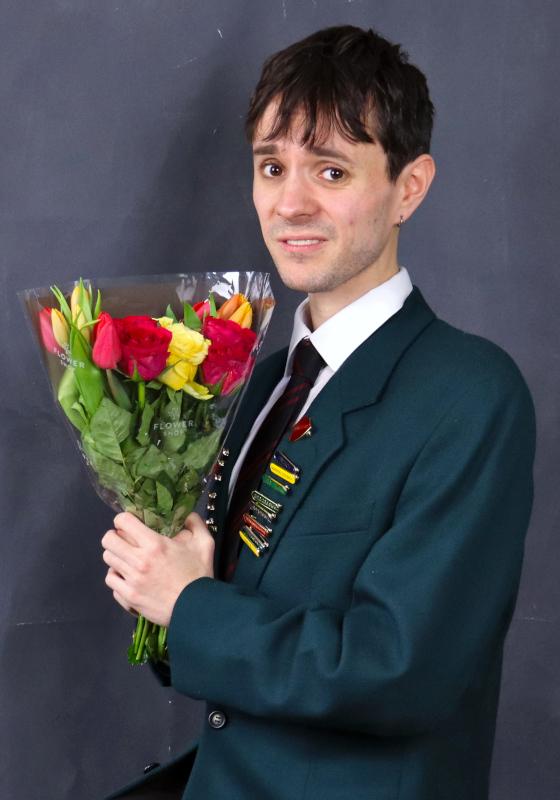 A head boy poses in a school blazer full of pin badges clutching a bunch of flowers and looking awkwardly at the viewer.