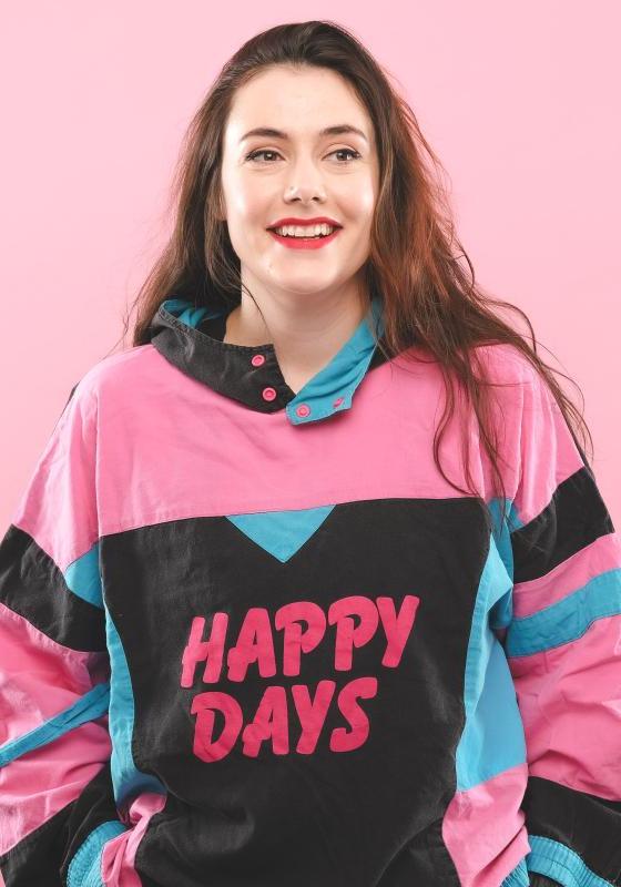 Image of Marjolein Robertson, performer, wearing a sweater that reads "HAPPY DAYS" in front of a pink background