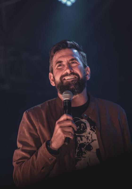 The performer is holding a microphone while standing under a stage spotlight