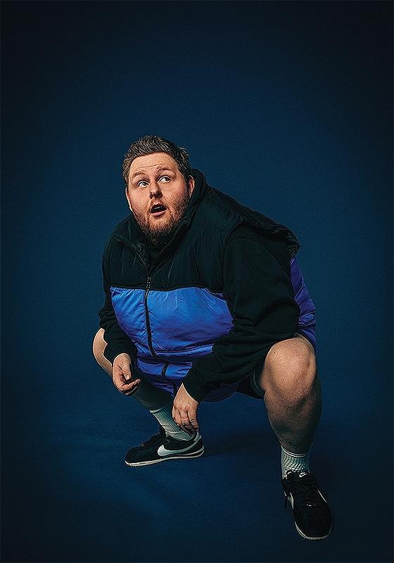 A large man squats down against a blue background wearing a blue puffer gilet, he looks agape up to the right corner of the image. 