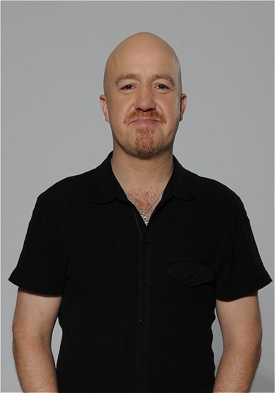 Andy Parsons stands facing the camera with a smug look on his face. He is stood against a grey background.