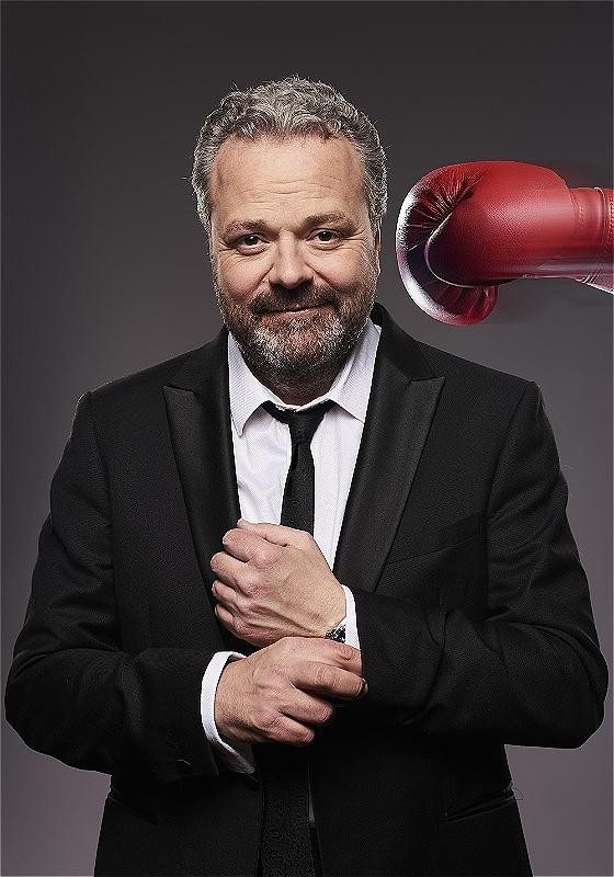 Hal faces the camera wearing a black suit, white shirt and black tie. He is doing up his cufflink. A red box glove comes from his right hand side about to hit his head. He is unaware of it.