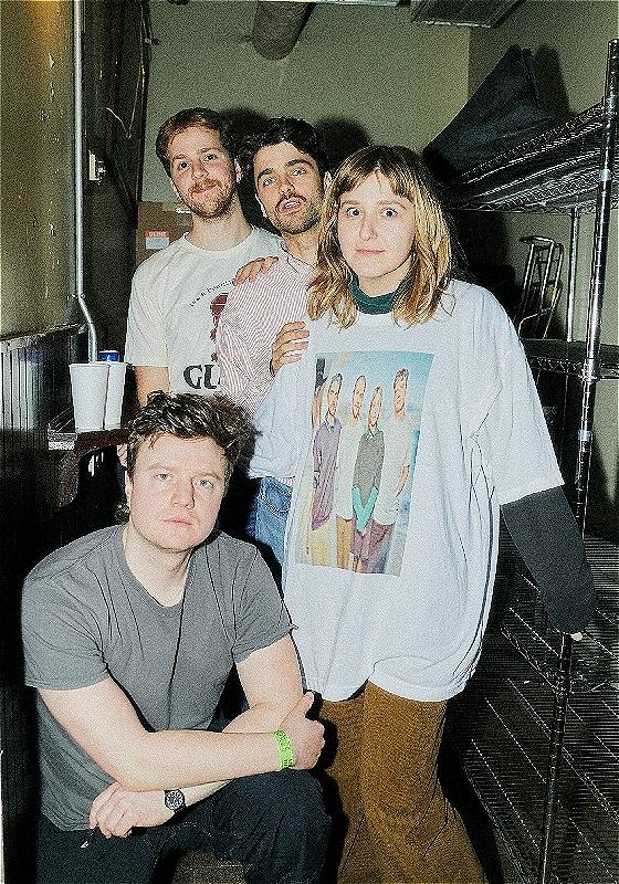 4 people stare at the camera. One is crouched on the floor in a grey t-shirt on the left hand side. The other three stand in a line on the right hand side. They stare startled into the camera. 