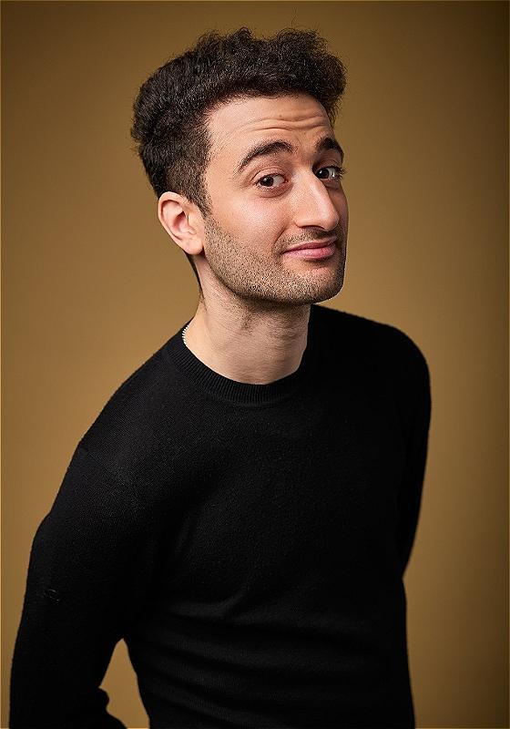 Hasan stands off centre to the camera. He is wearing a black high neck shirt against a brown background. He looks at the camera with a confident expression. 