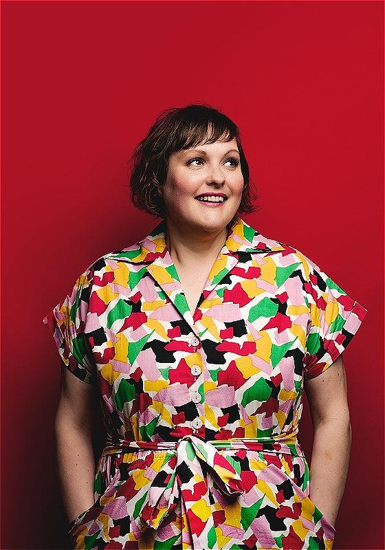 Josie Long in a colourful jumpsuit, with hands in pockets stares smilingly off to the right against a red background. 