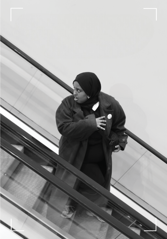 A black and white image of the performer running down an escalator, holding her bag and looking over her shoulder in fear.