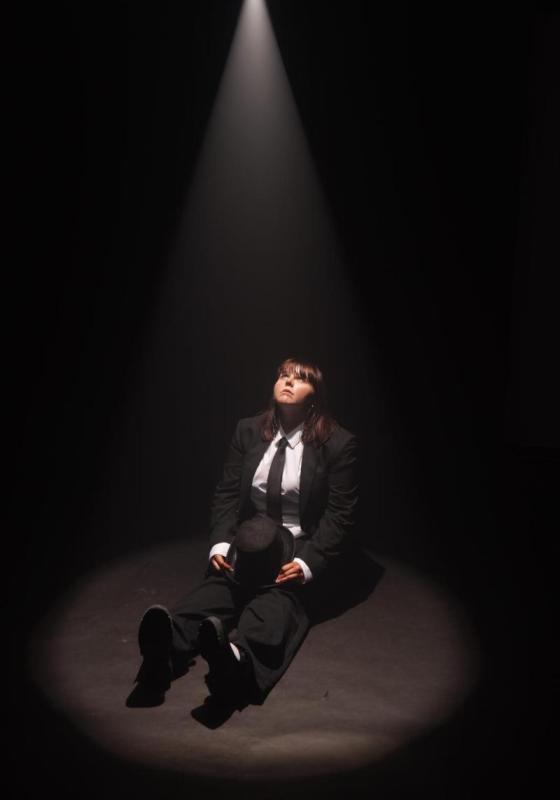 A performer dressed in a black suit and tie sits on the floor, a spotlight shining on them. Their legs are stretched out in front of them, they're holding a hat on their lap, and they are staring up at the spotlight.
