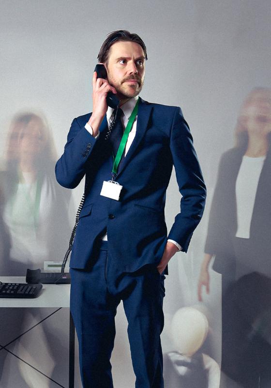 Man in suit wearing lanyard holds phone to ear looking pensively across blurry background with screaming office folk.