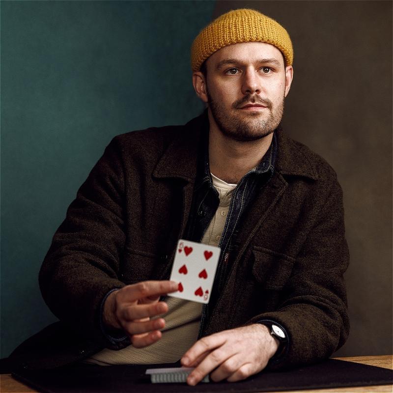The performer is in front of a dark background, wearing a yellow hat and brown jacket. They hold a playing card in their hand. 