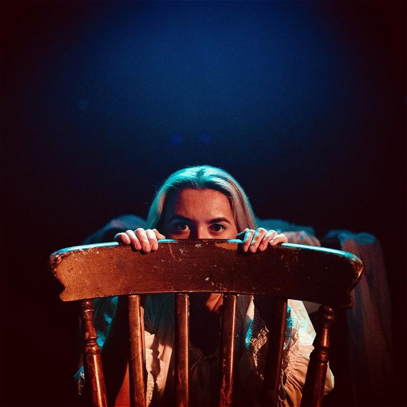 A woman with blonde hair peeking over the top of a wooden chair, her eyes visible as she grips the chair's back. The scene is lit with a warm, theatrical blue light, creating a moody and mysterious atmosphere.