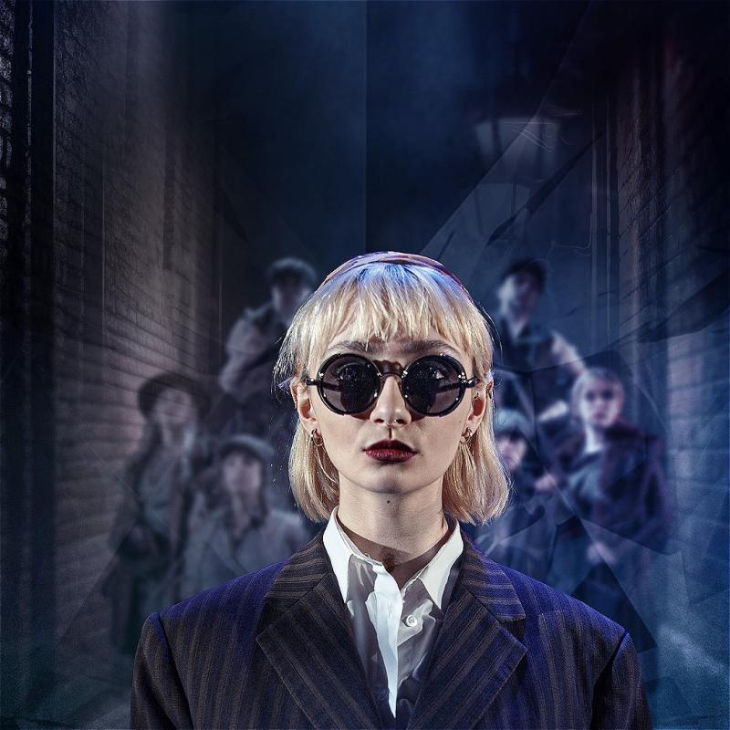 Woman in sunglasses and a dark suit stands looks into the camera. There is a group of blurred figures in Victorian style clothing behind her in a dimly lit alley.