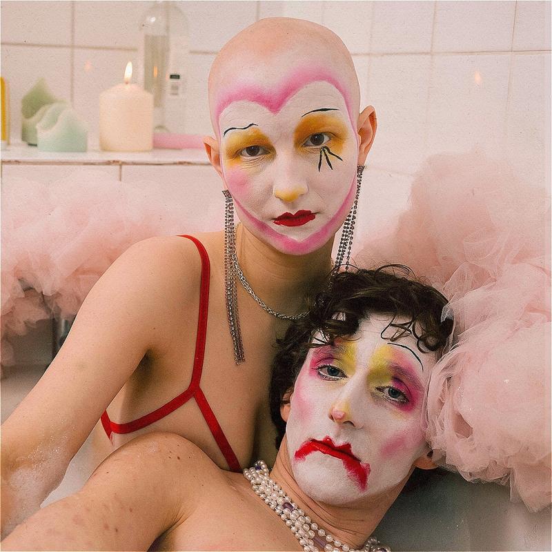 Two people pose in a bathtub with dramatic clown-style makeup. Around the bath there is pink tulle and a candle is lit.