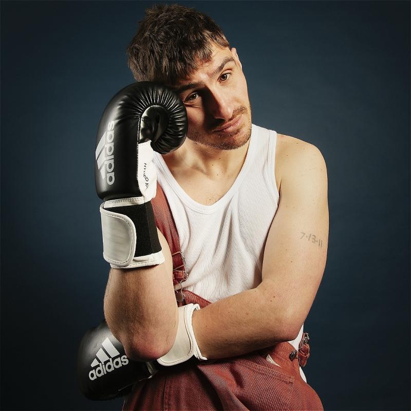 Young man with a contemplative expression wearing boxing gloves and a white tank top, leaning his cheek on one glove against a blue background.