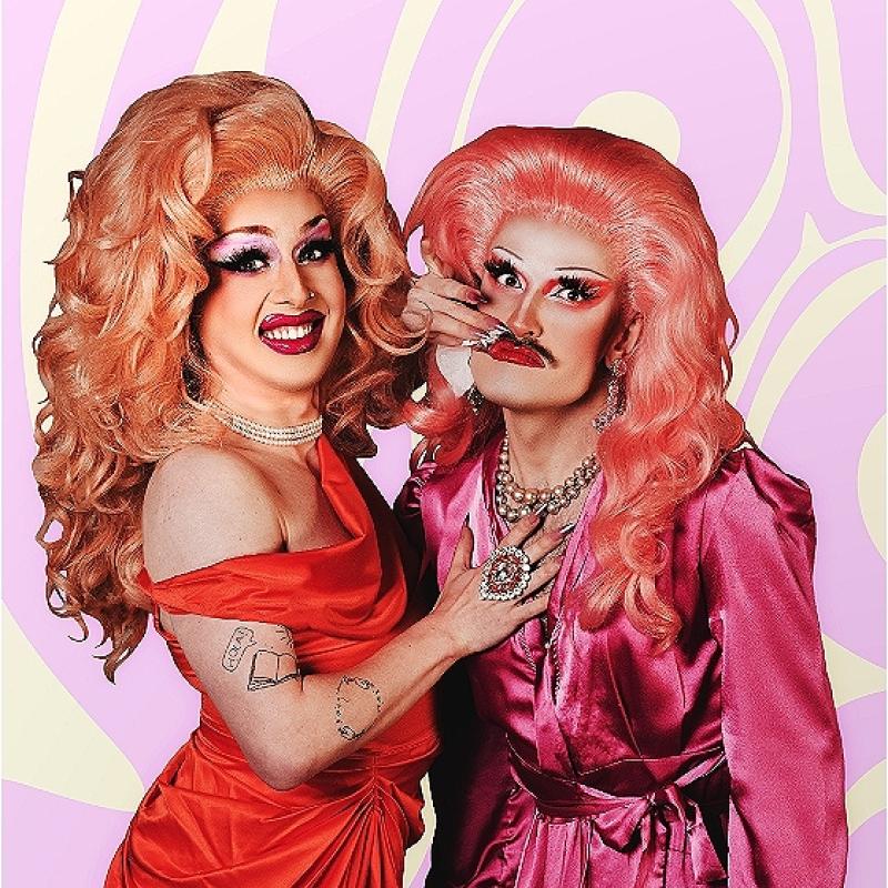 Two drag artists, sporting pink hair and vibrant makeup, looking nervously ahead.