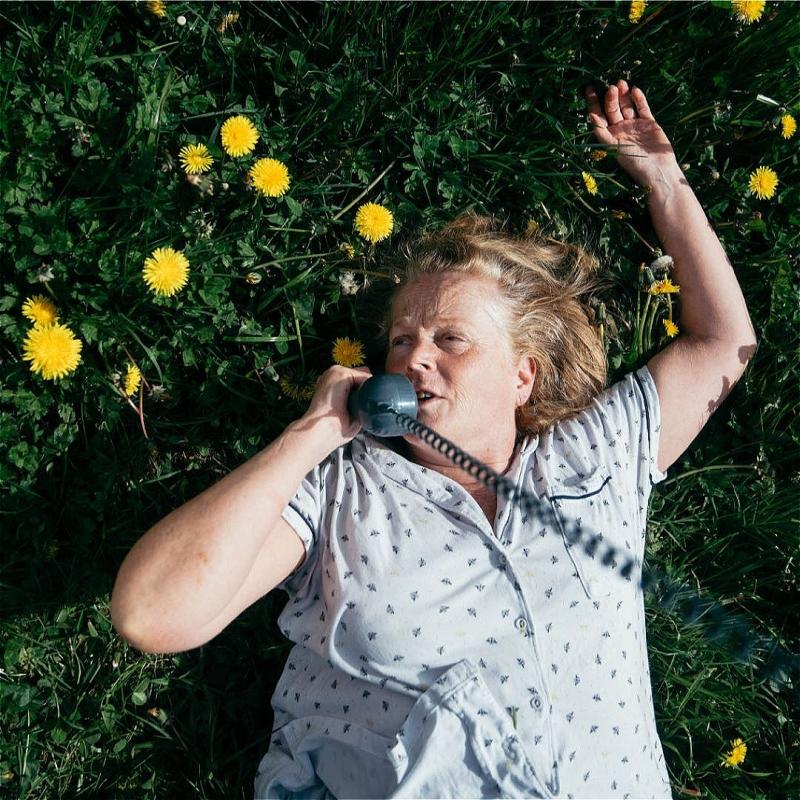 Woman lying on her back in a field of grass and dandelions, talking on an old-fashioned rotary phone.