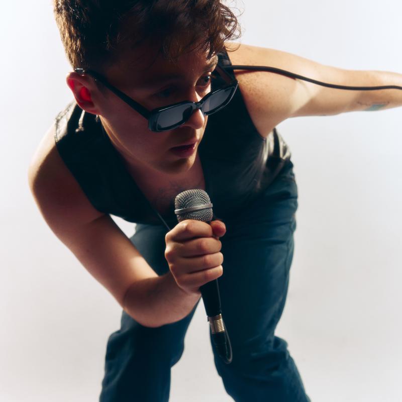 Jake Roche, arms bare in a waistcoat, jeans, and stylish sunglasses, crouches with a microphone in hand. Jake is lit from the side, and the background is white. 