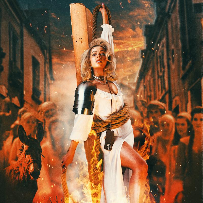 Abi wearing white shirt and shoulder armour is tied to a plank of wood with sparks of fire surrounding her. Blurred silhouettes of historical figures make up a crowd behind her.