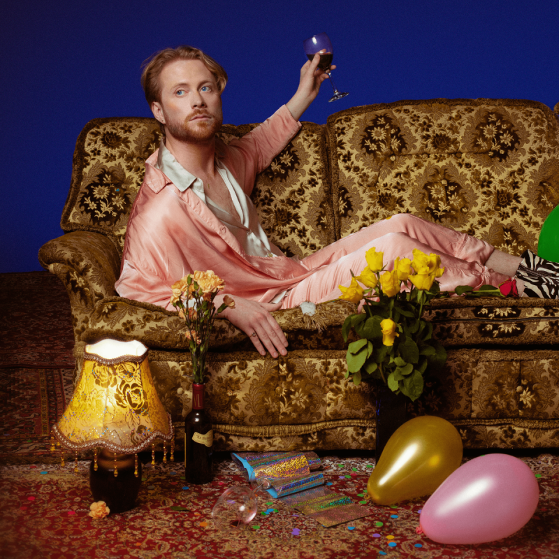 Performer sitting on gold sofa holding wine glass wearing pink silk pjs surrounded by balloons, flowers, wine and lamp