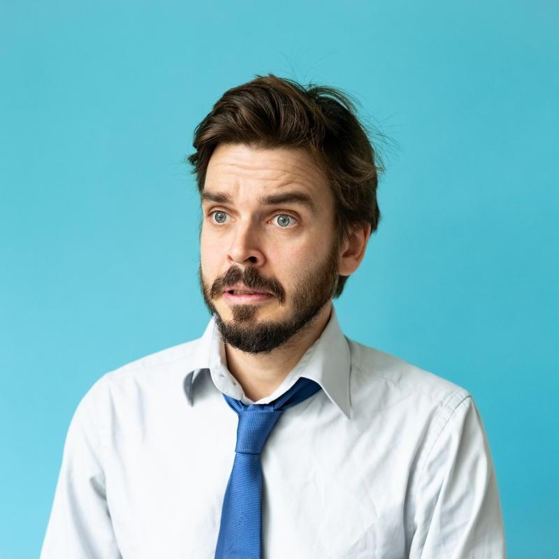 Bearded man in white shirt and blue tie looking puzzled in front of a blue backdrop.