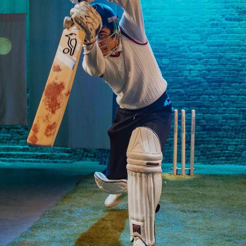 A man on stage in cricketing attire swings a cricket bat towards the audience.