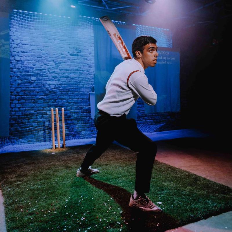 A man on stage in cricketing attire swings a cricket bat towards the audience.