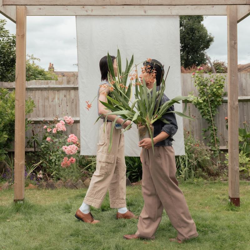 In a garden, Ghost and John are carrying large house plants as they meet in the middle under a wooden arch frame.