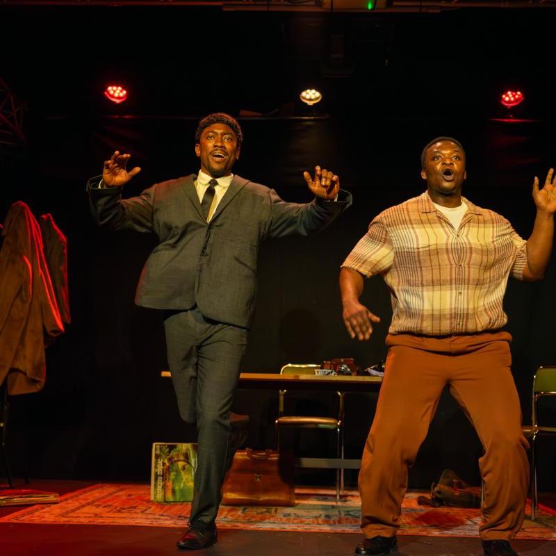 Two actors on stage with hands raised in a theatrical performance, under warm stage lighting.