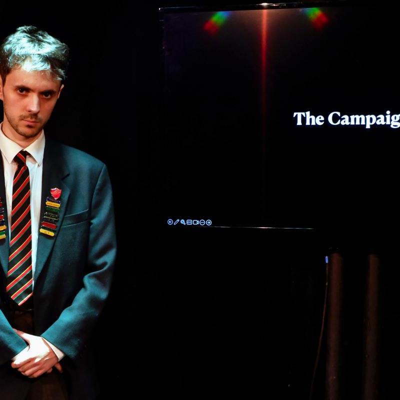 School boy left of a projected image that says 'The Campaign' looking directly at the camera