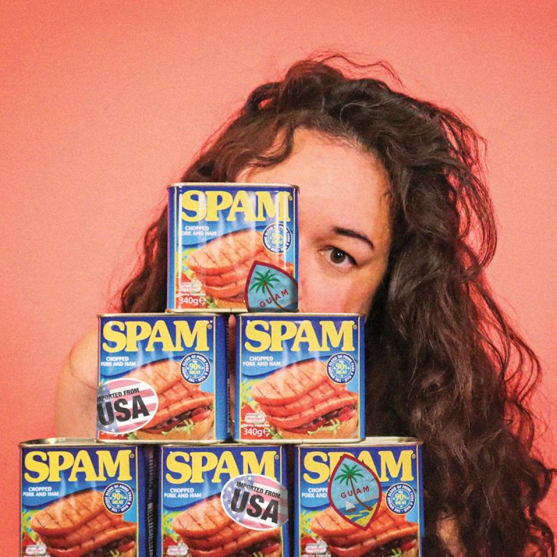 Curly-haired individual standing behind a stack of SPAM canned meat products against a coral background.