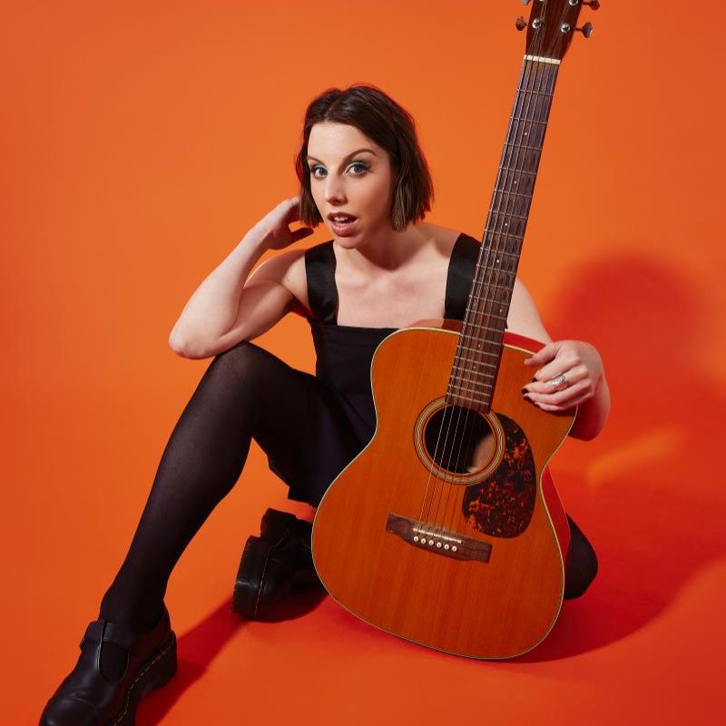 A person sat holding a guitar against an orange background.