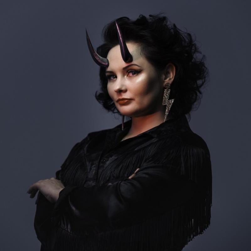 Person with black-fringed outfit, devil horns and lightning bolt earring against a grey background.