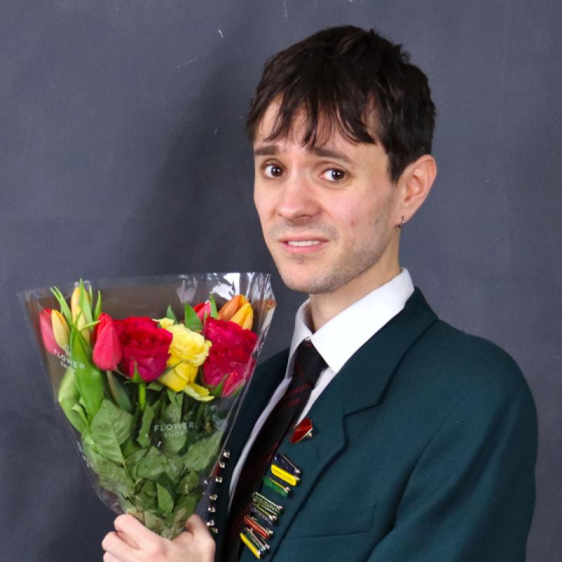 A head boy poses in a school blazer full of pin badges clutching a bunch of flowers and looking awkwardly at the viewer.