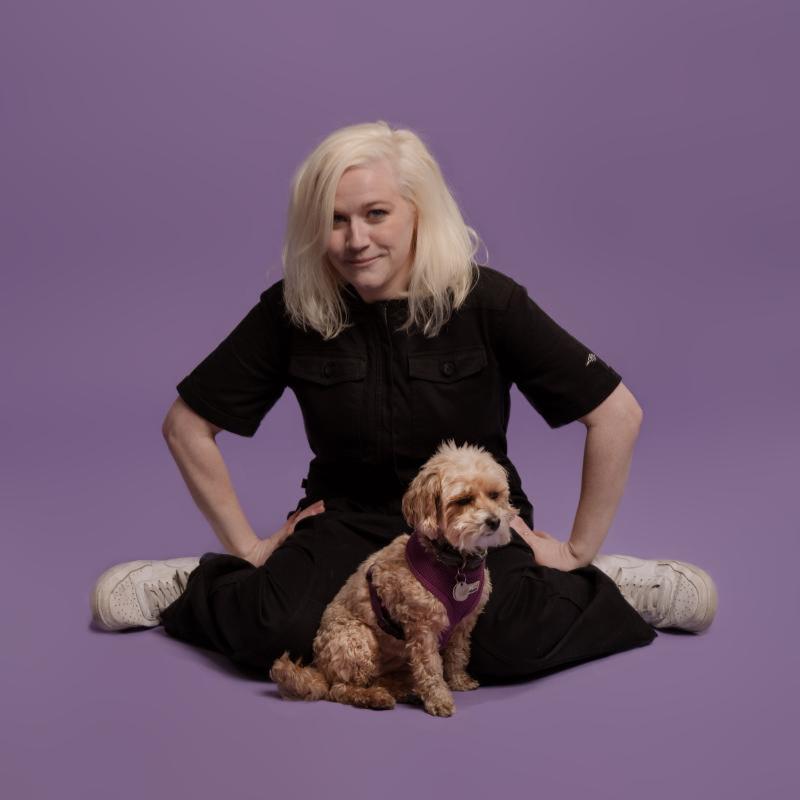 Photo of the performer wearing a black jumpsuit and trainers against a lilac background. Their small dog sits directly in front of them.