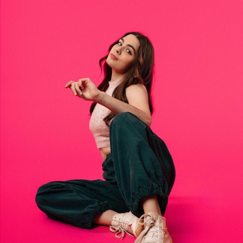 Photo of Amelia against a bright pink background wearing a pastel pink tank top, emerald cord trousers. and white trainers.