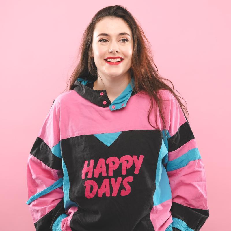 Image of Marjolein Robertson, performer, wearing a sweater that reads "HAPPY DAYS" in front of a pink background