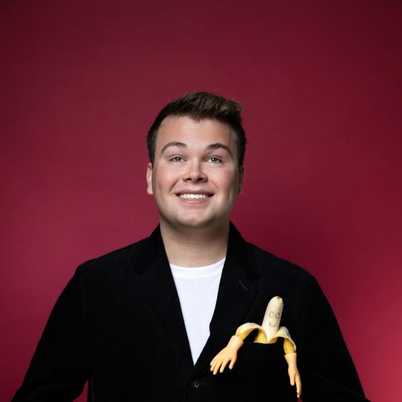 Image of Max Fulham, performer, with a banana with a doodled-on face in his cardigan pocket in front of a red background, the banana peels have small hands