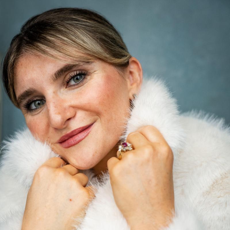 Sophie smiles at the camera wearing a cozy white fluffy jumper and a sparkly ring. Their hands are snuggled up towards their chin.