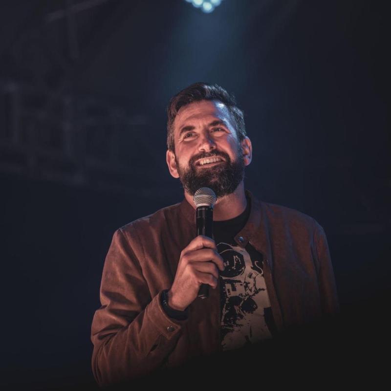 The performer is holding a microphone while standing under a stage spotlight