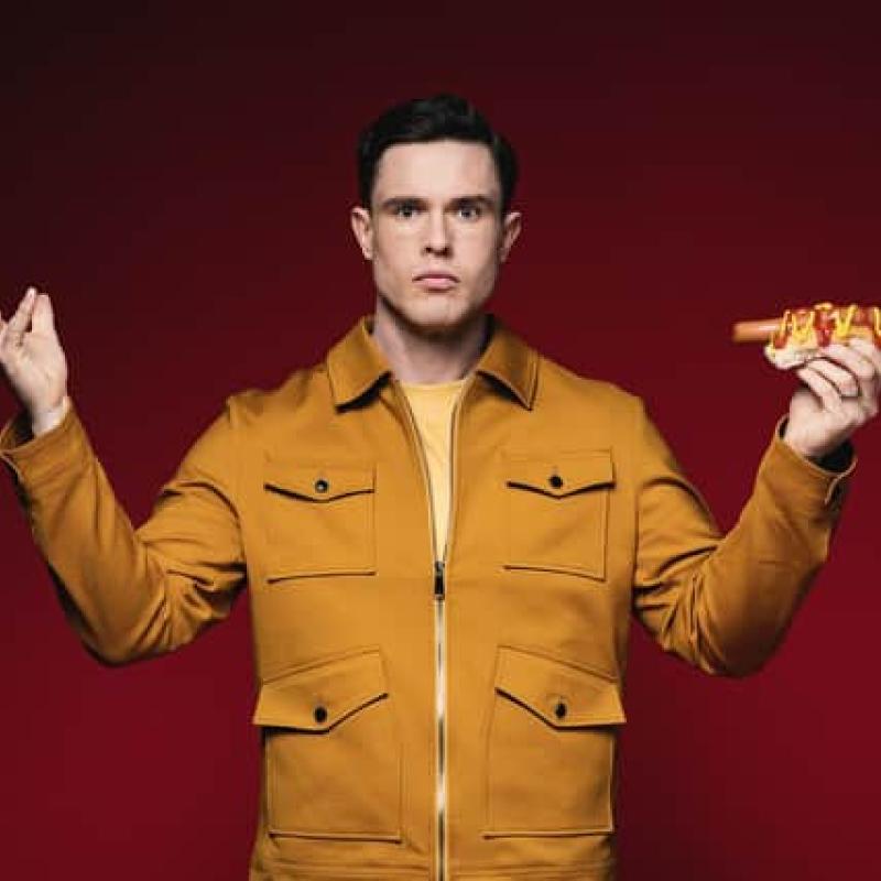 Ed Gamble looks out to the camera holding a hot dog in their left hand.