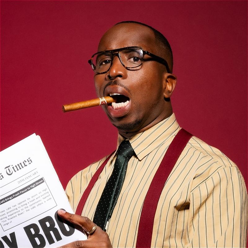 Kiell Smith-Bynoe looks to the camera with black glasses on and a cigar in his mouth. He wears a pink shirt with thin black stripes, a black tie and burgundy braces. He holds a newspaper in his hand. 