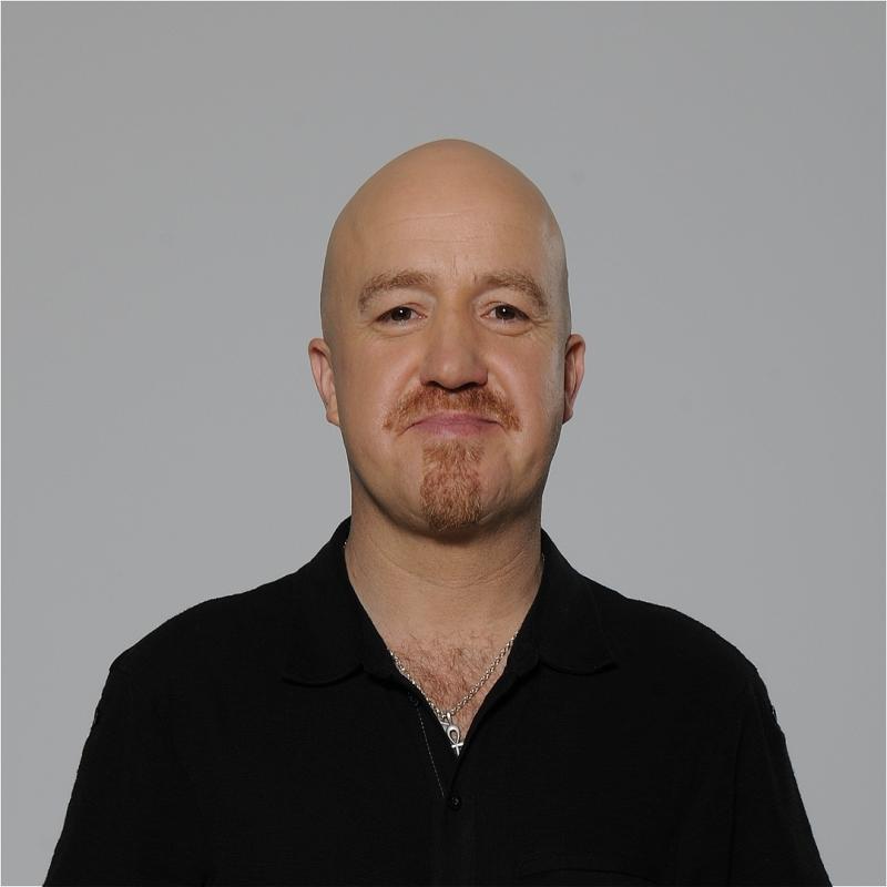 Andy Parsons stands facing the camera with a smug look on his face. He is stood against a grey background.