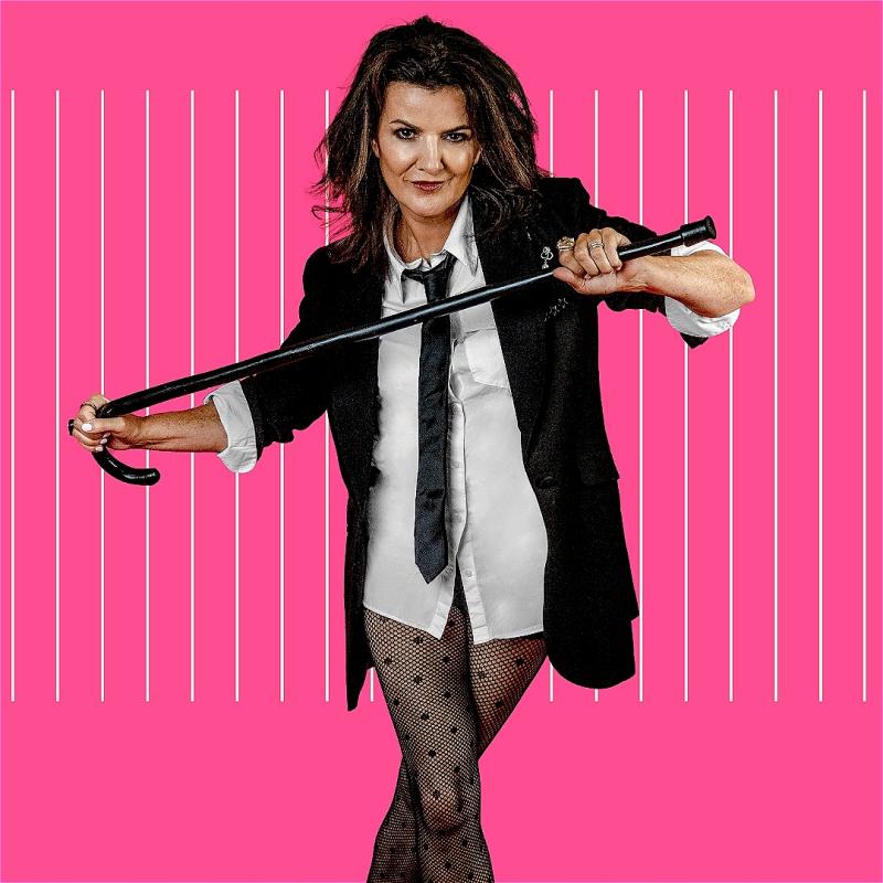 Deirdre stands facing the camera against a hot pink background. They are wearing a black blazer, white shirt, black tie and fishnet tights. They hold a black kane in their hand. They have a seductive look on their face.