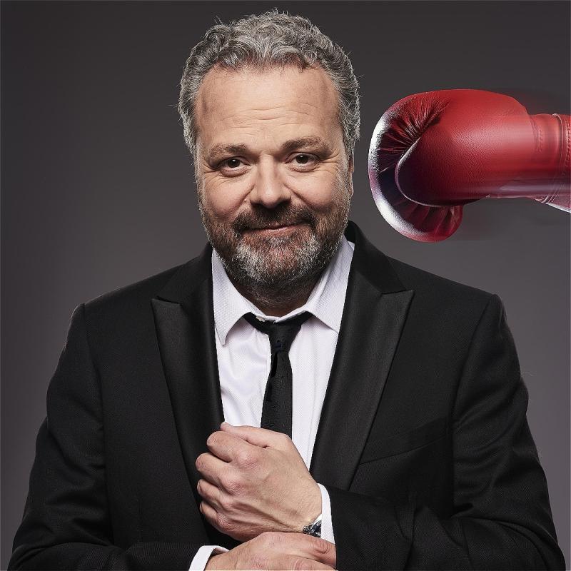 Hal faces the camera wearing a black suit, white shirt and black tie. He is doing up his cufflink. A red box glove comes from his right hand side about to hit his head. He is unaware of it.