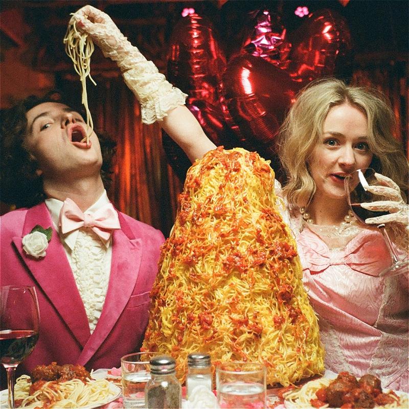 A woman feeds a man a bit of spaghetti from her hand as she sips red wine. There is a mountain of spaghetti in front of her. It has an 80s wedding vibe about it, a lot of pink. 