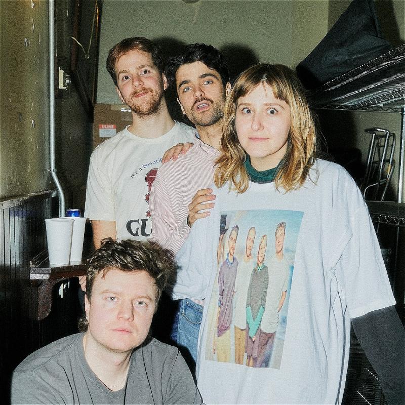 4 people stare at the camera. One is crouched on the floor in a grey t-shirt on the left hand side. The other three stand in a line on the right hand side. They stare startled into the camera. 