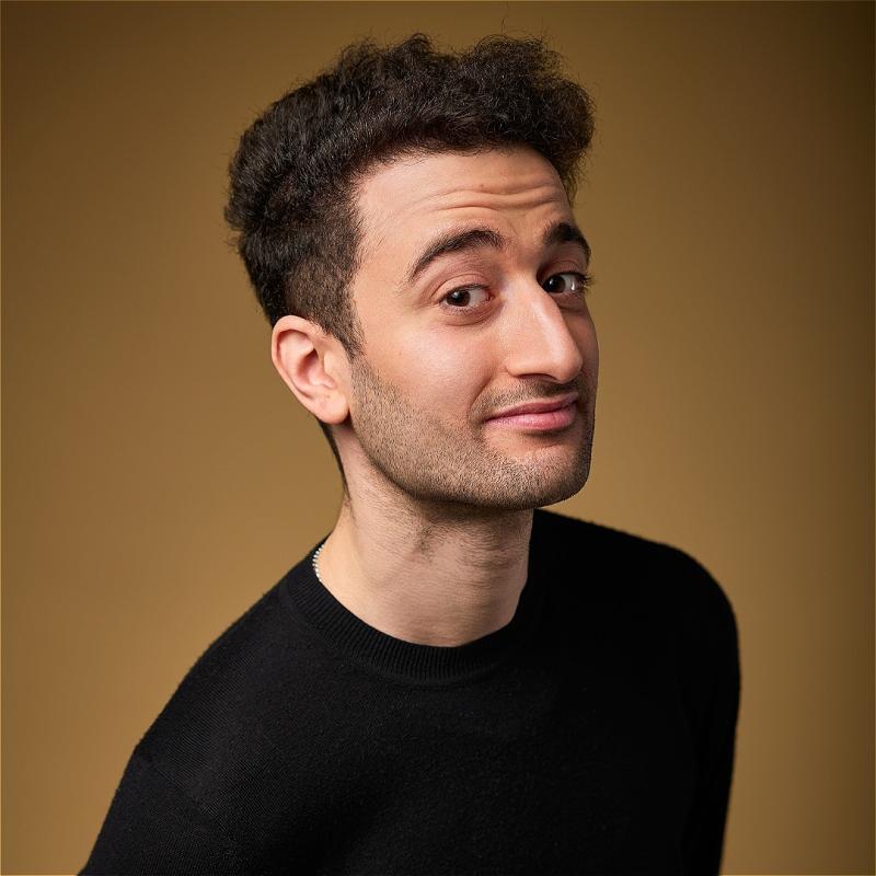 Hasan stands off centre to the camera. He is wearing a black high neck shirt against a brown background. He looks at the camera with a confident expression. 