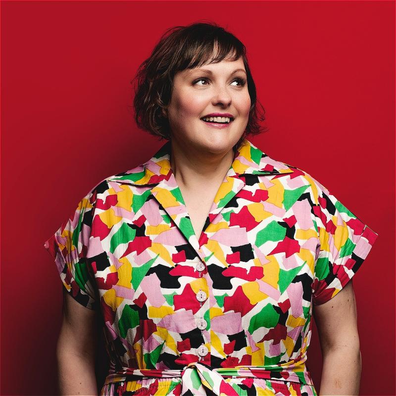 Josie Long in a colourful jumpsuit, with hands in pockets stares smilingly off to the right against a red background. 