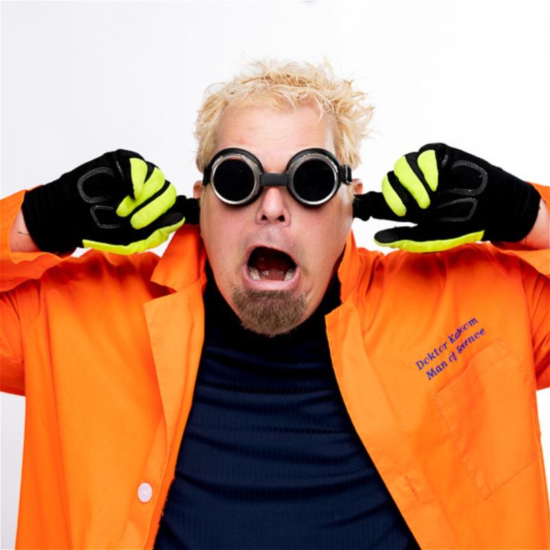 Man wearing black goggles, and gloves in an orange lab coat. Stands with his mouth agape in shock and his fingers in his ears, directly looking into the camera.